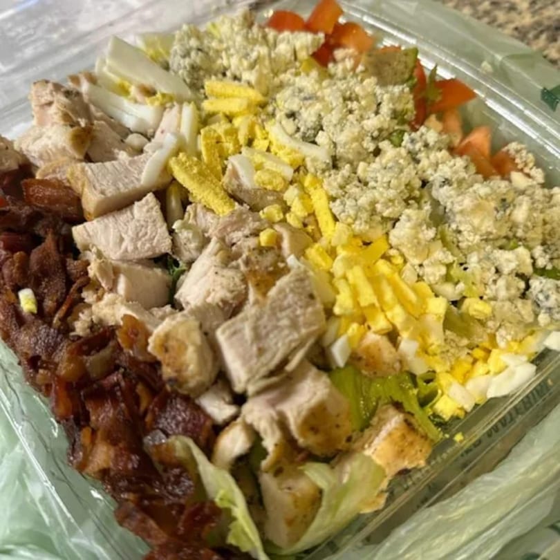 A close-up of a vibrant Cobb salad in a plastic container, featuring chicken, bacon, eggs, and blue cheese.